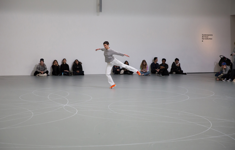 A dancer in white pants, a grey top and orange sneakers hovers on one foot. Her other leg is stretched behind her parallel to the floor. People are seated against the wall and watch.